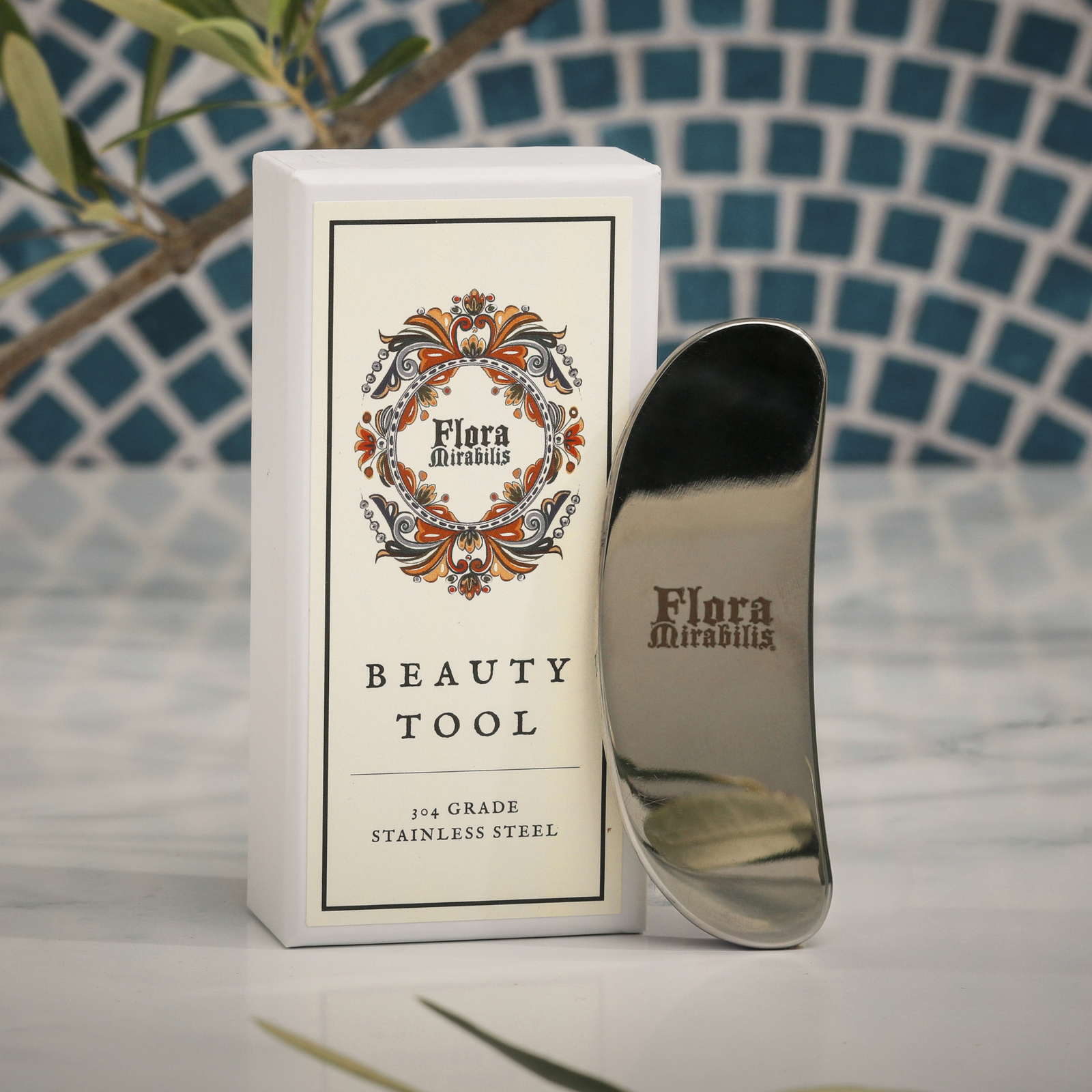 Flora Mirabilis stainless steel Beauty Tool displayed upright beside its decorative packaging on a marble surface.