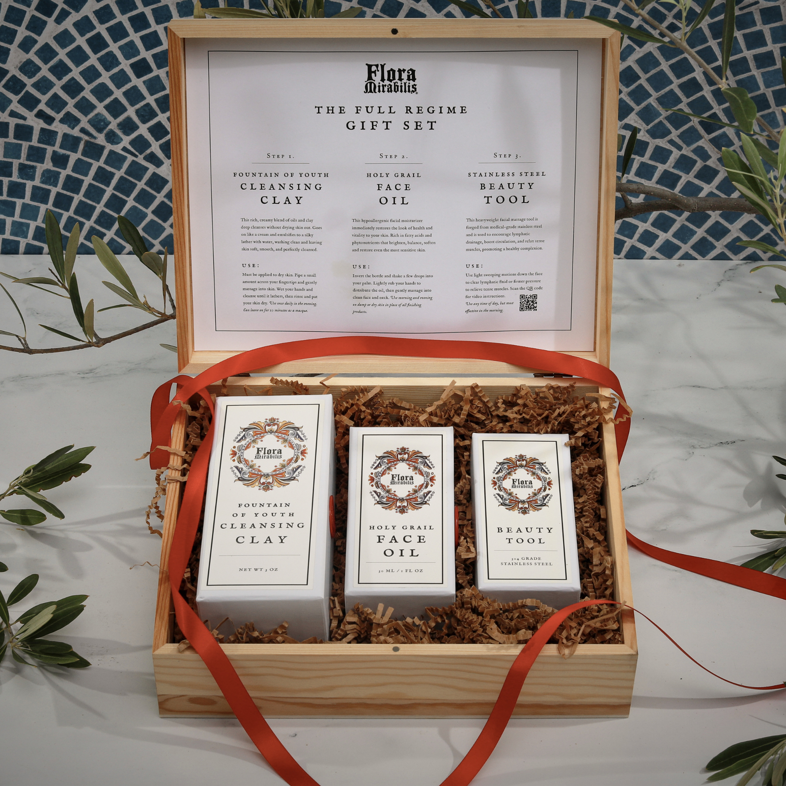 Flora Mirabilis Full Regime Gift Set displayed in an open wooden box with the Cleansing Clay, Holy Grail Face Oil, and Beauty Tool arranged inside with red ribbon and kraft filler.