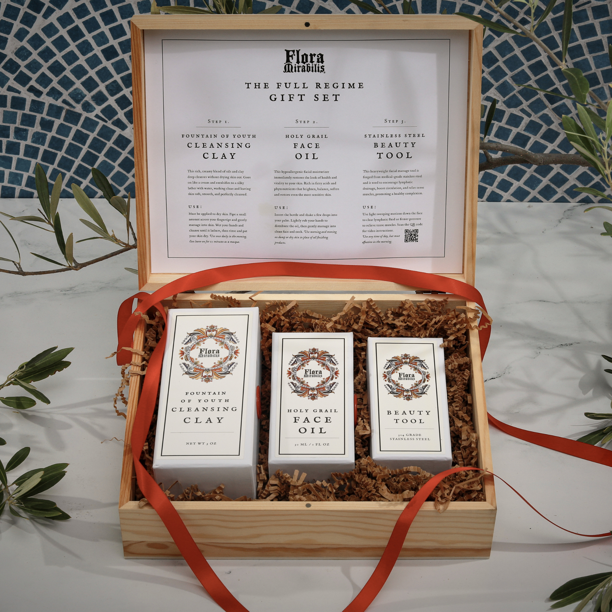 Flora Mirabilis Full Regime Gift Set displayed in an open wooden box with the Cleansing Clay, Holy Grail Face Oil, and Beauty Tool arranged inside with red ribbon and kraft filler.