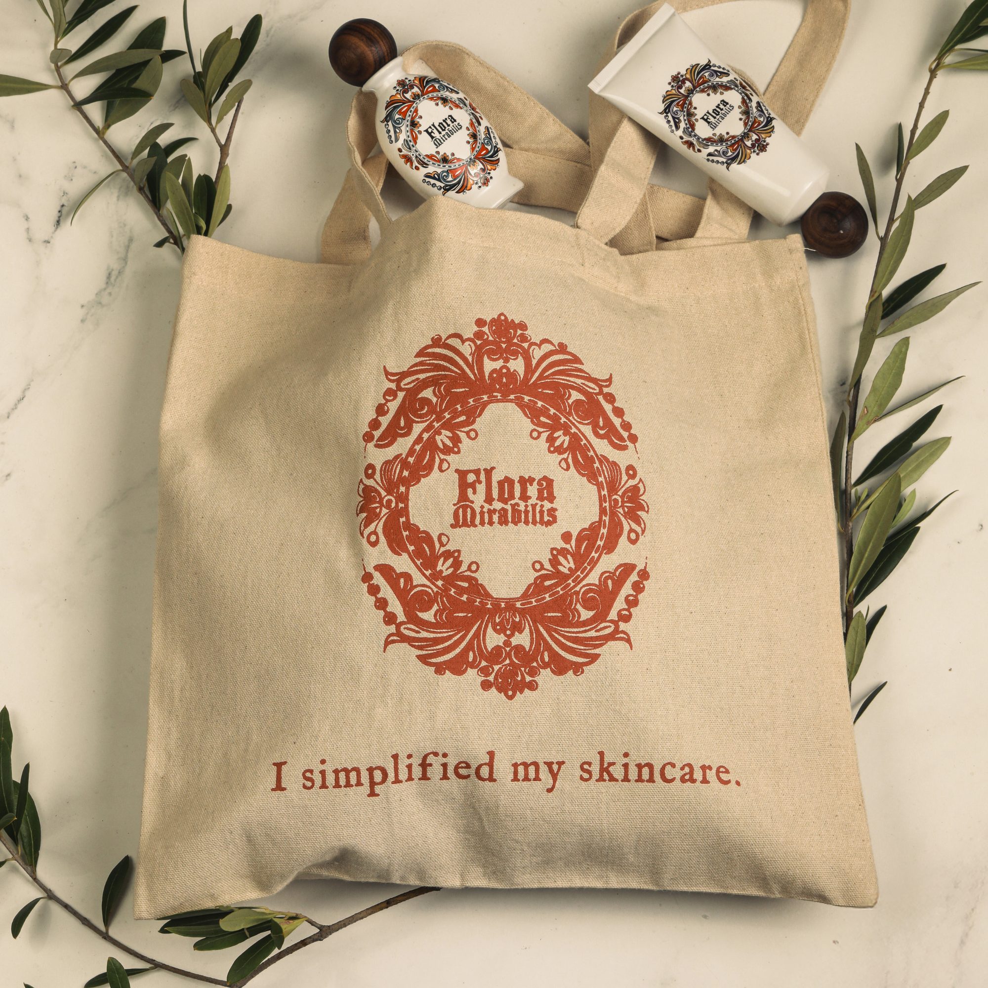 Flora Mirabilis canvas tote bag with red logo and ‘I simplified my skincare’ text, displayed with Holy Grail Face Oil and Cleansing Clay on a marble surface with olive branches.