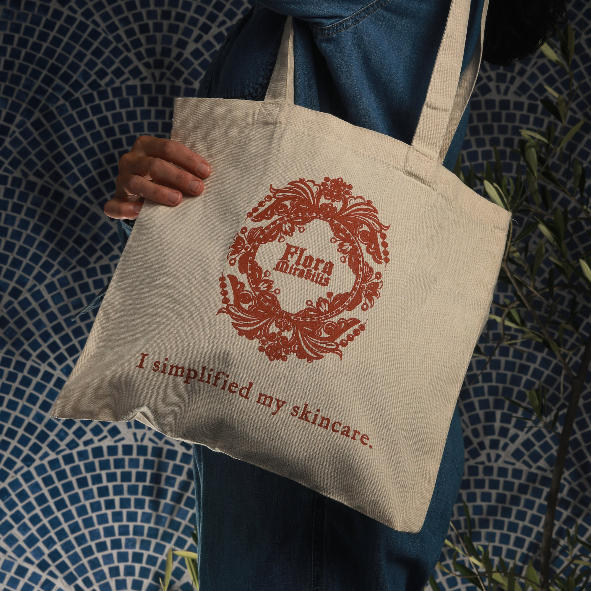 Person holding a Flora Mirabilis canvas tote bag with red logo and ‘I simplified my skincare’ text against a blue mosaic background.