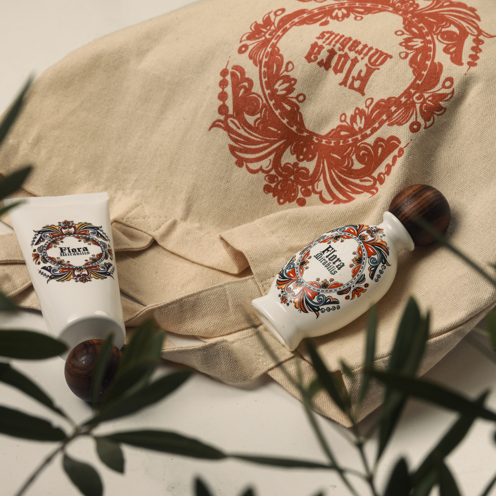 Flora Mirabilis canvas tote bag with red logo surrounded by Holy Grail Face Oil and Cleansing Clay tubes, styled with olive branches.