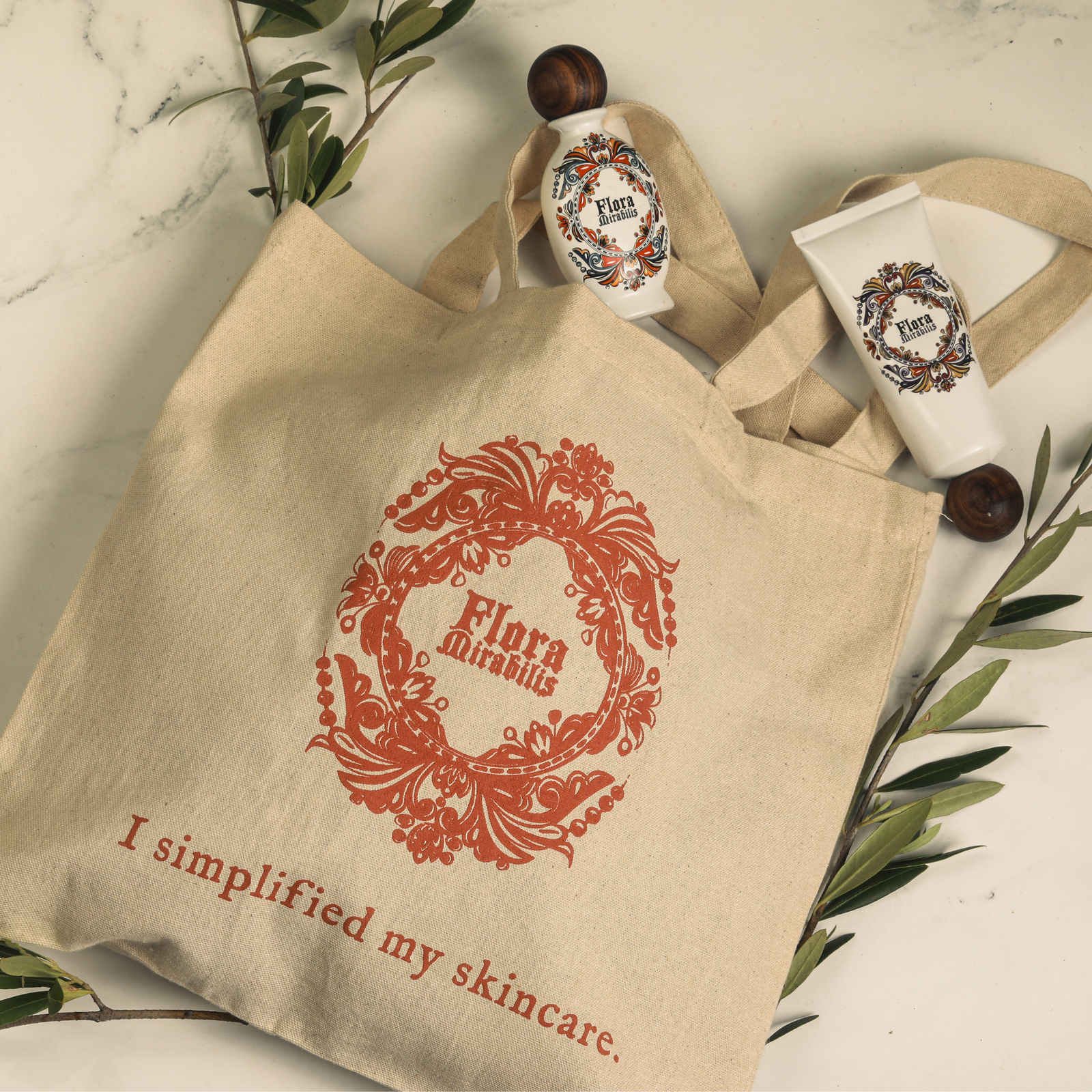 Overhead view of Flora Mirabilis canvas tote bag with red logo and ‘I simplified my skincare’ text, styled with Holy Grail Face Oil, Cleansing Clay, and olive branches.