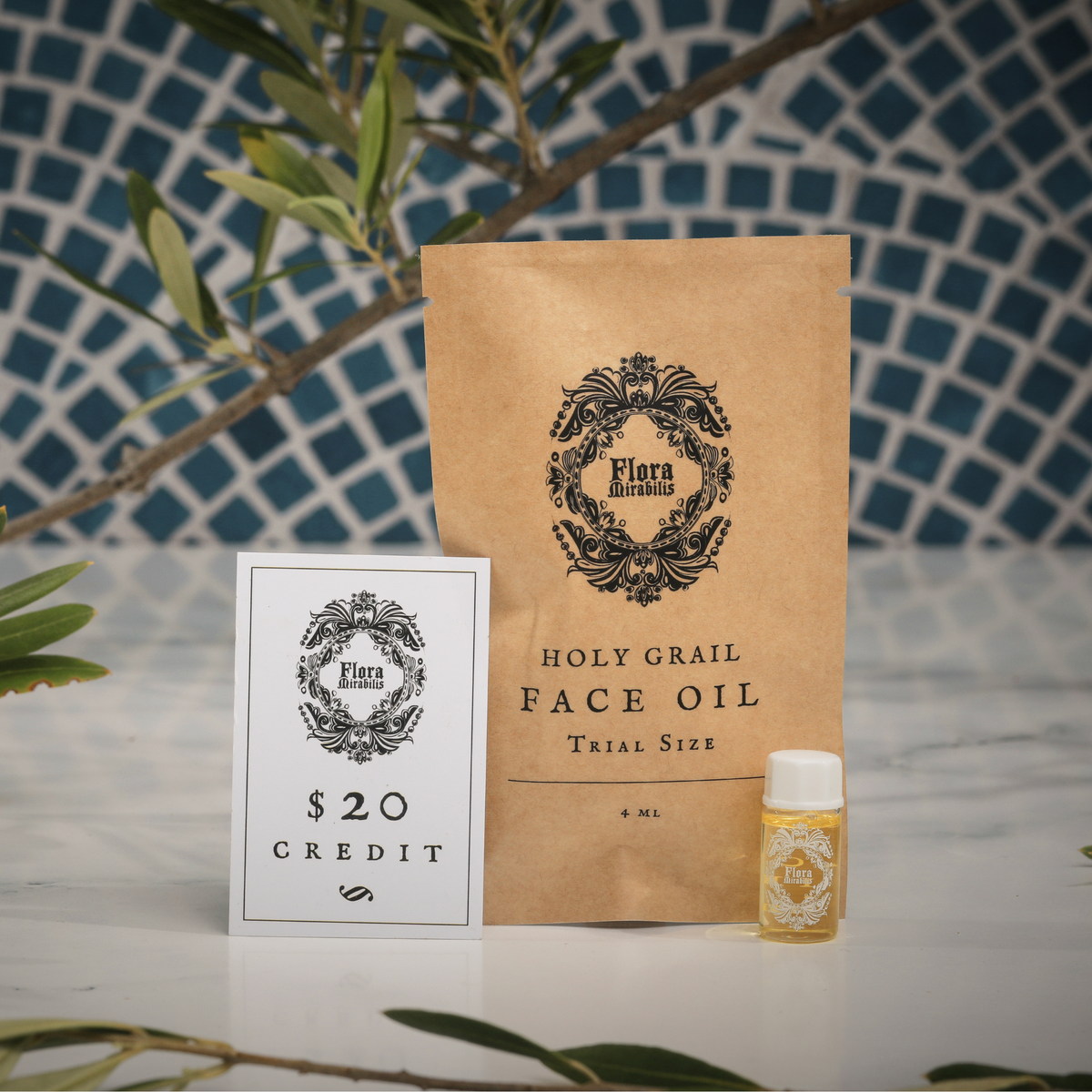 Flora Mirabilis Holy Grail Face Oil trial-size kit displayed with pouch, $20 credit card insert, and mini vial on a marble surface with mosaic tile background.
